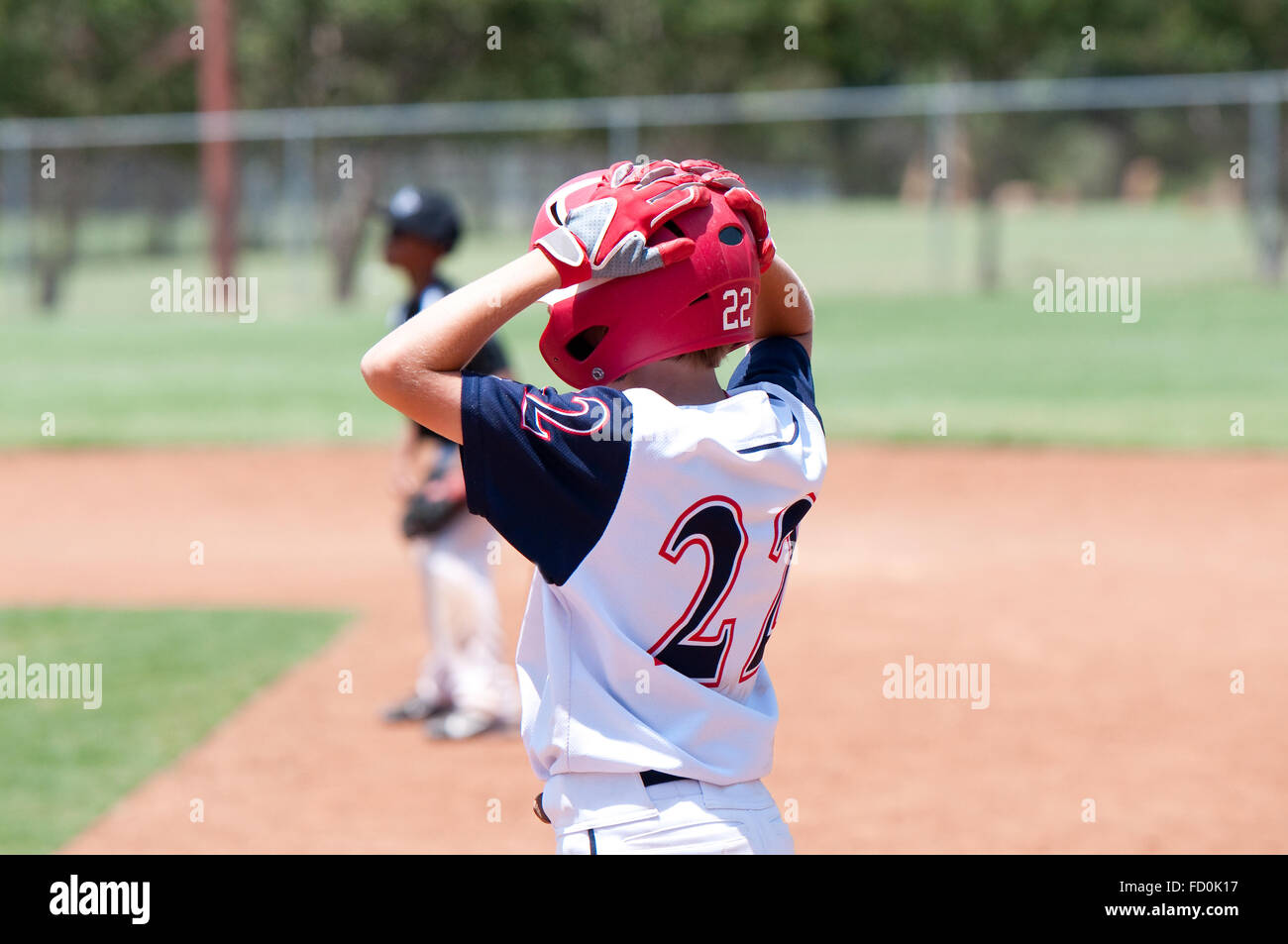 Youth baseball player wearing red helmet standing on first base with hands on head. Stock Photo