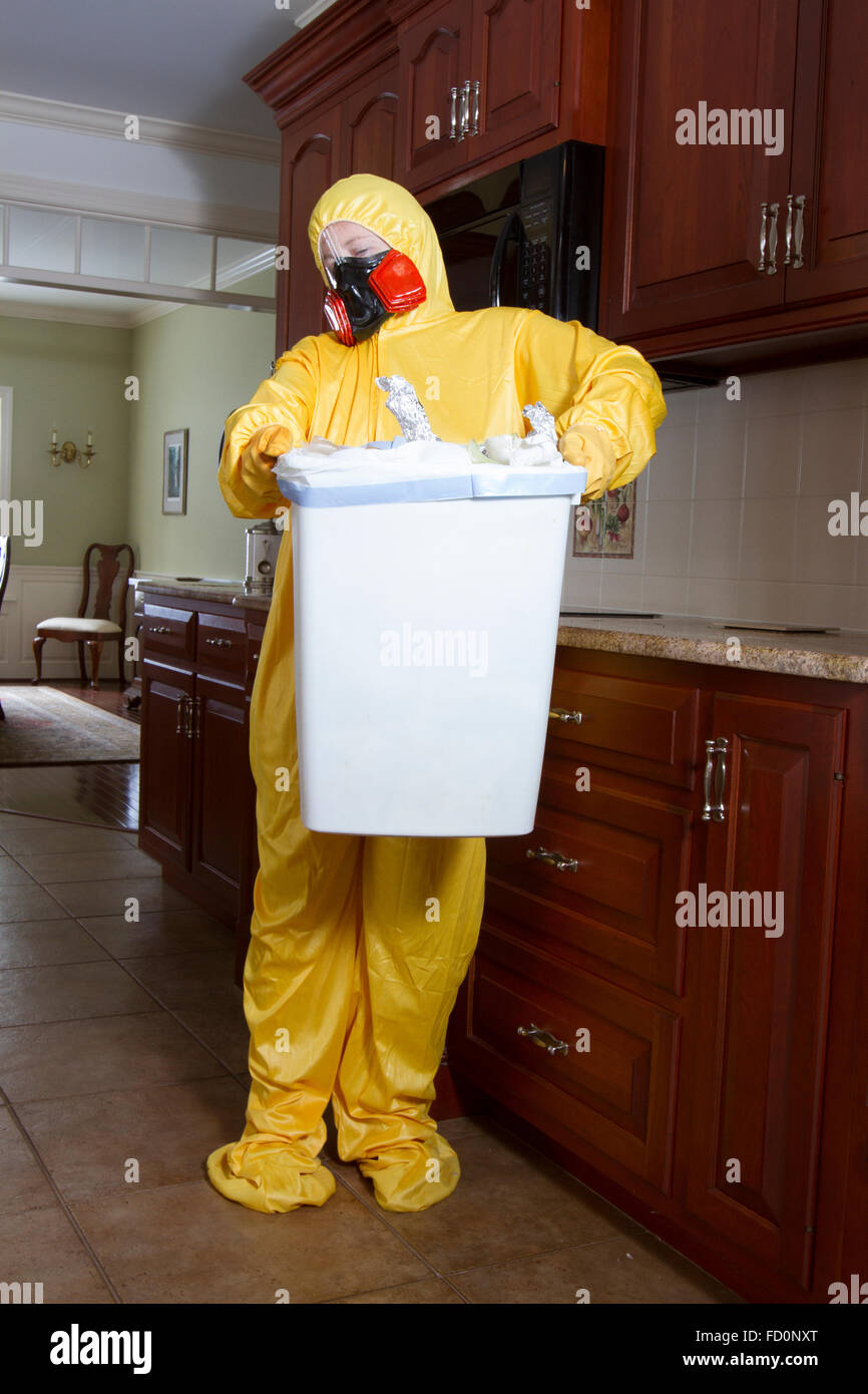 Woman dressed in yellow haz mat outfit with ventilator taking out the garbage in kitchen. Stock Photo