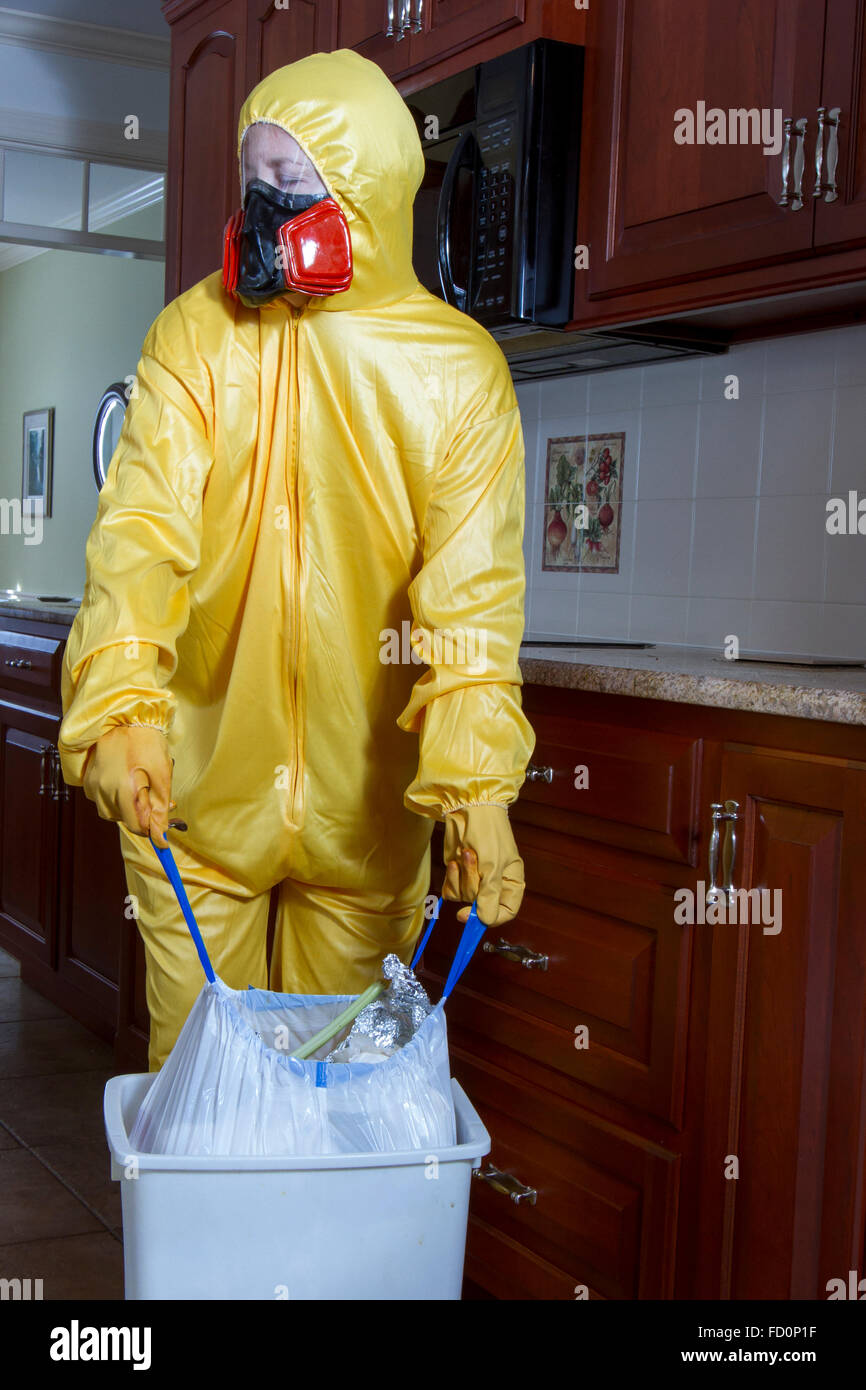 Woman dressed in yellow haz mat outfit with ventilator taking out the garbage in kitchen. Stock Photo