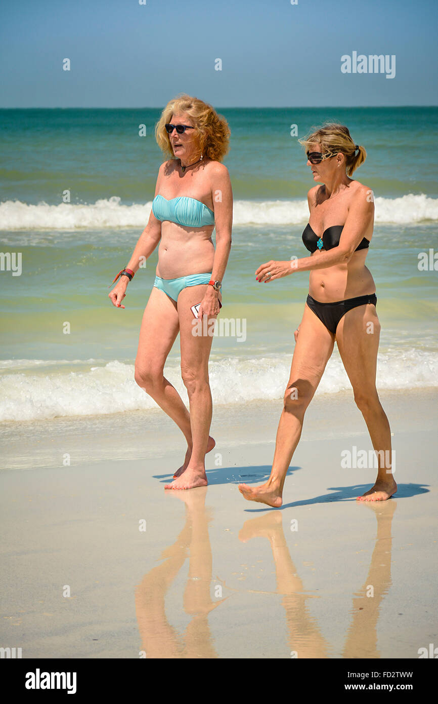 Two older middle aged women in bikinis stroll the beach conversing and enjoying the white sand and the nearby gentle waves Stock Photo