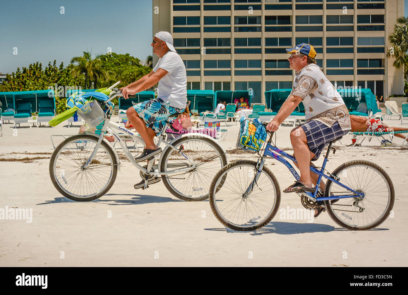 Two men loaded with beach gear bike along the shoreline on their cruiser bicycles in front of sunbathers and condos in Florida Stock Photo