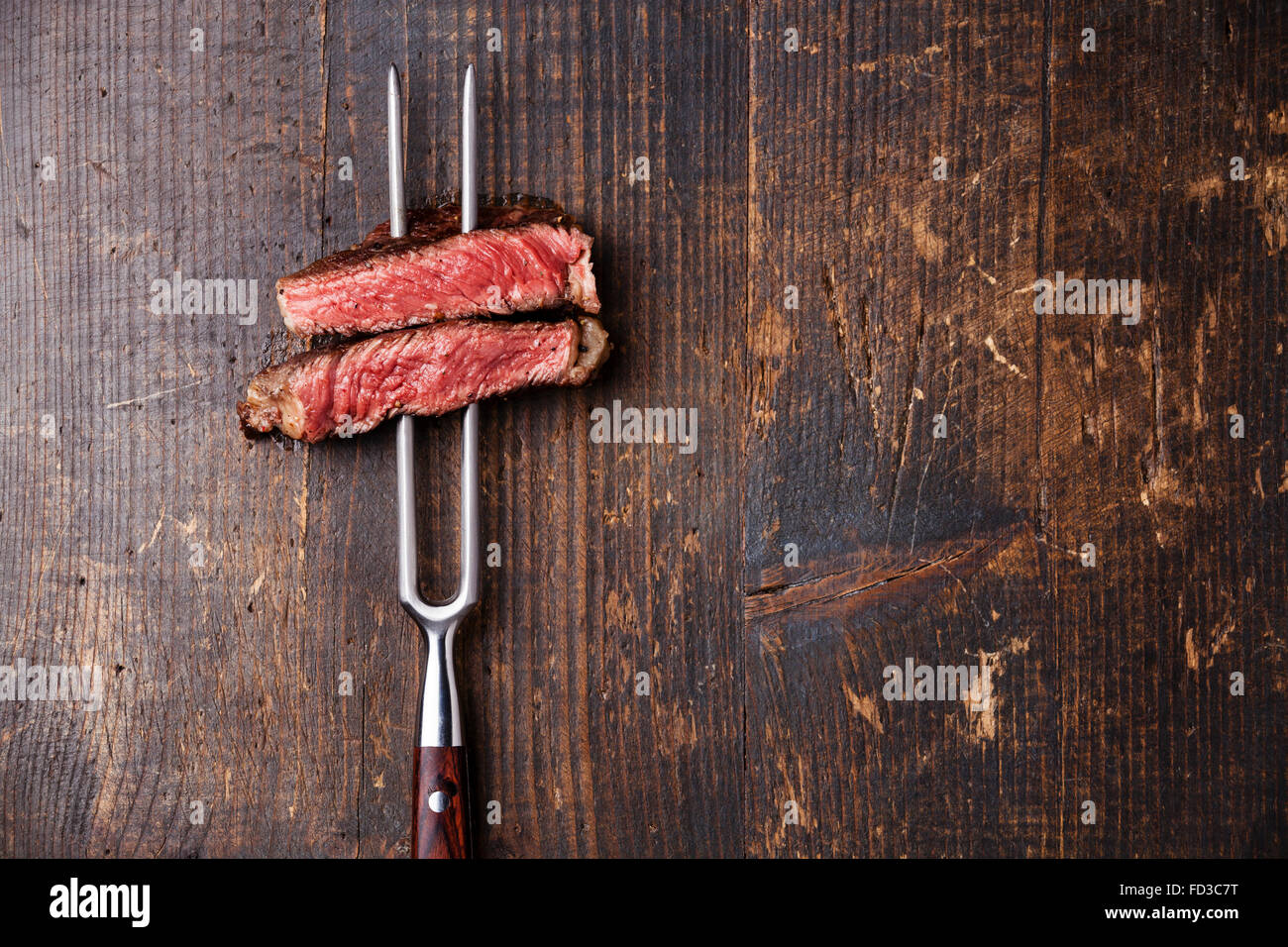 Slices of Medium rare grilled Steak Ribeye on meat fork on dark wooden