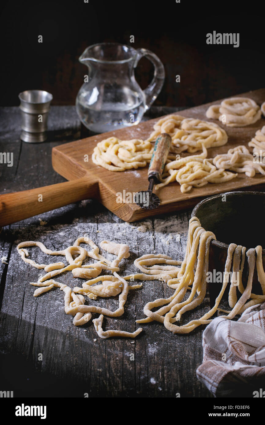 Fresh homemade pici pasta on wood chopping board over old wooden table with flour, copper bowl, rolling-pin and galss jug of wat Stock Photo