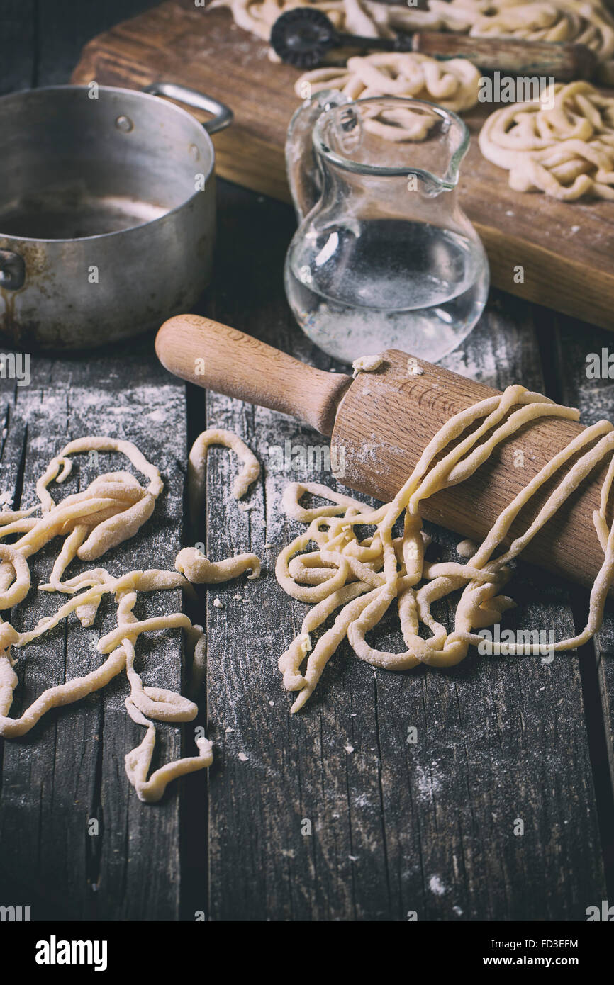 Fresh homemade pici pasta on wood chopping board over old wooden table with flour, aluminum pan, rolling-pin and galss jug of wa Stock Photo