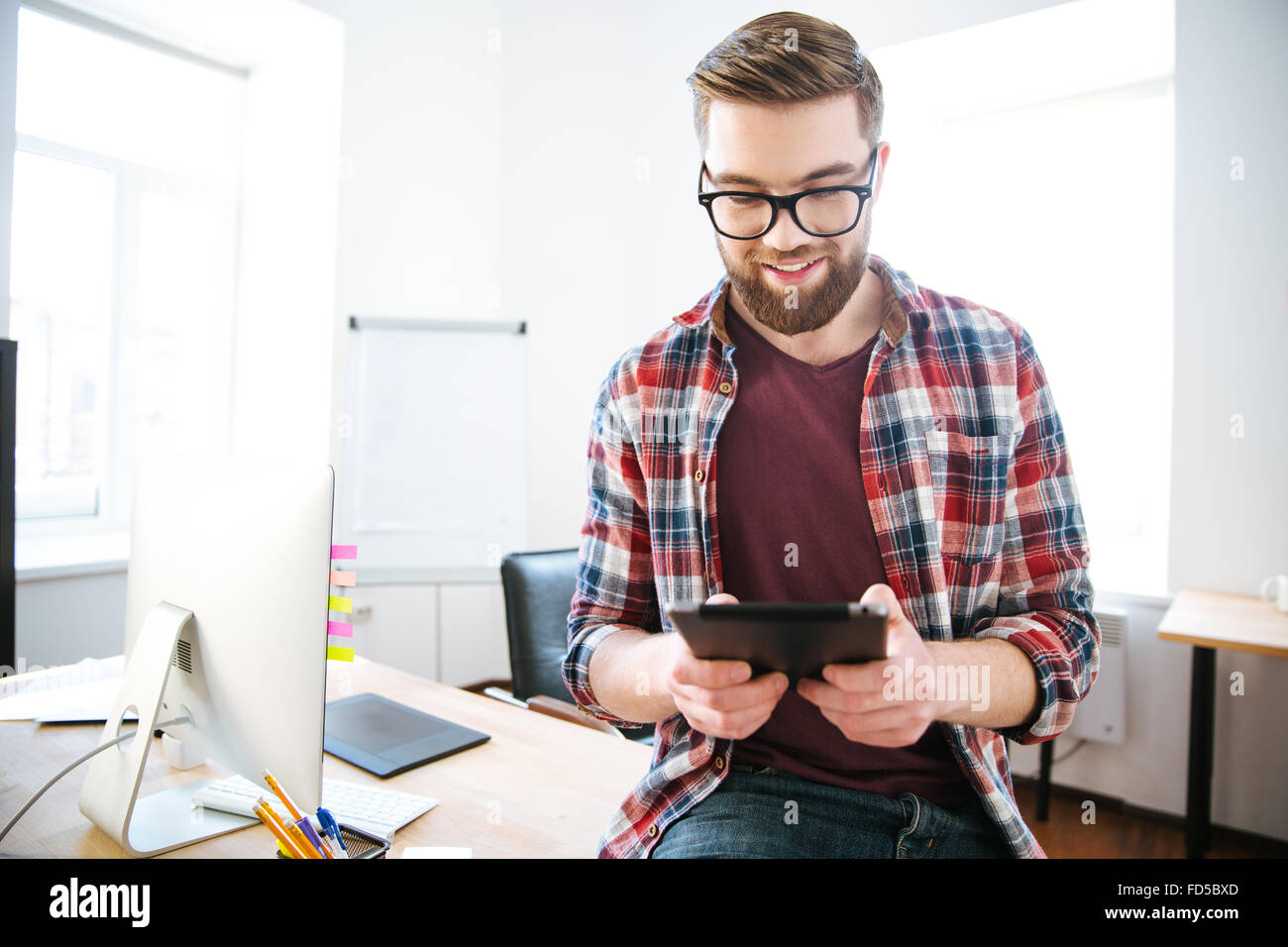 Happy handsome man with beard in plaid shirt and glasses sitting on the table in office and using tablet Stock Photo