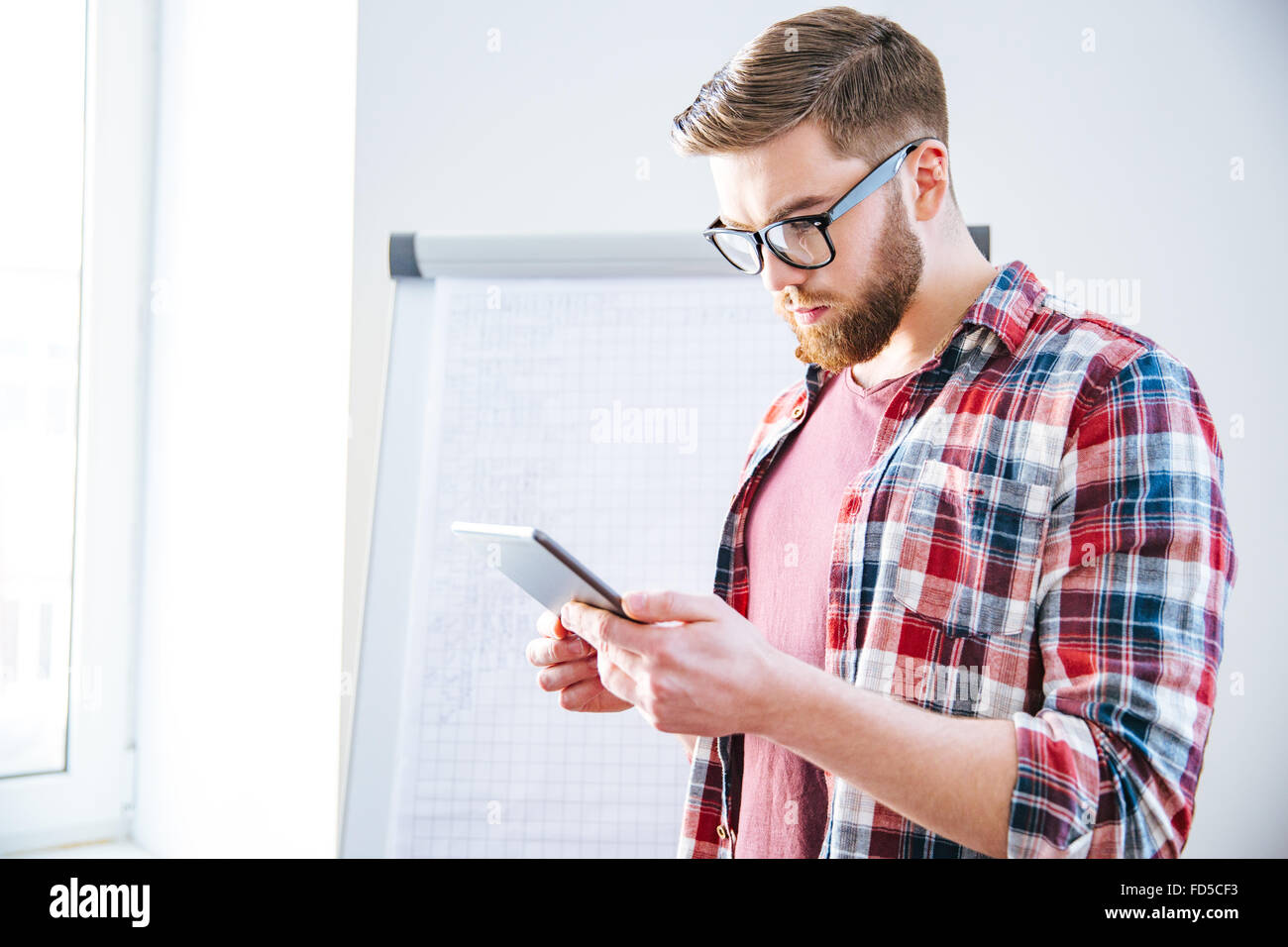 Handsome serious young man with beard in plaid shirt standing near flipchart and using tablet Stock Photo