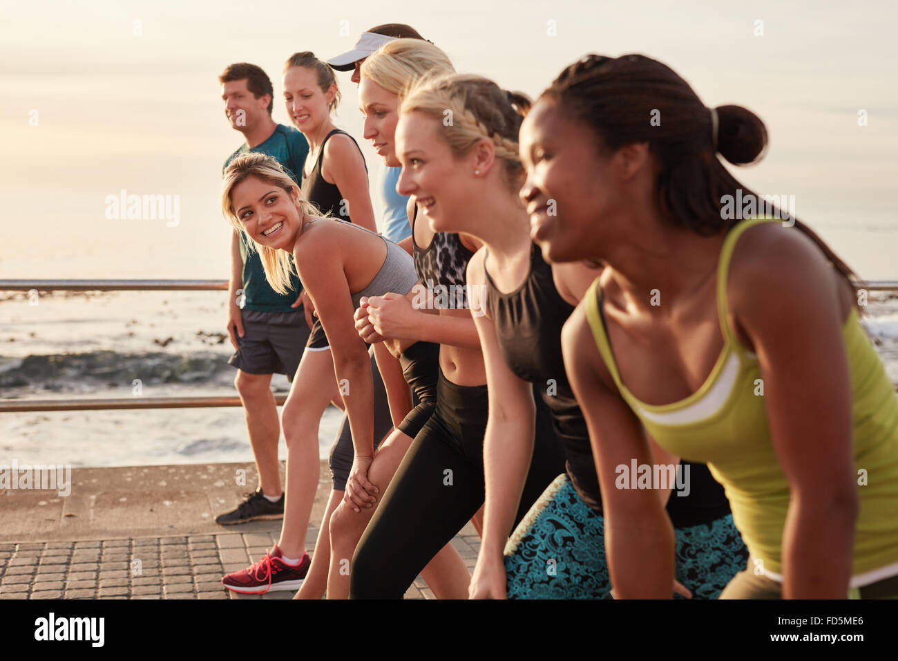 Group of young athletes in start position, focus on woman. Fit young people preparing for race along sea. Stock Photo