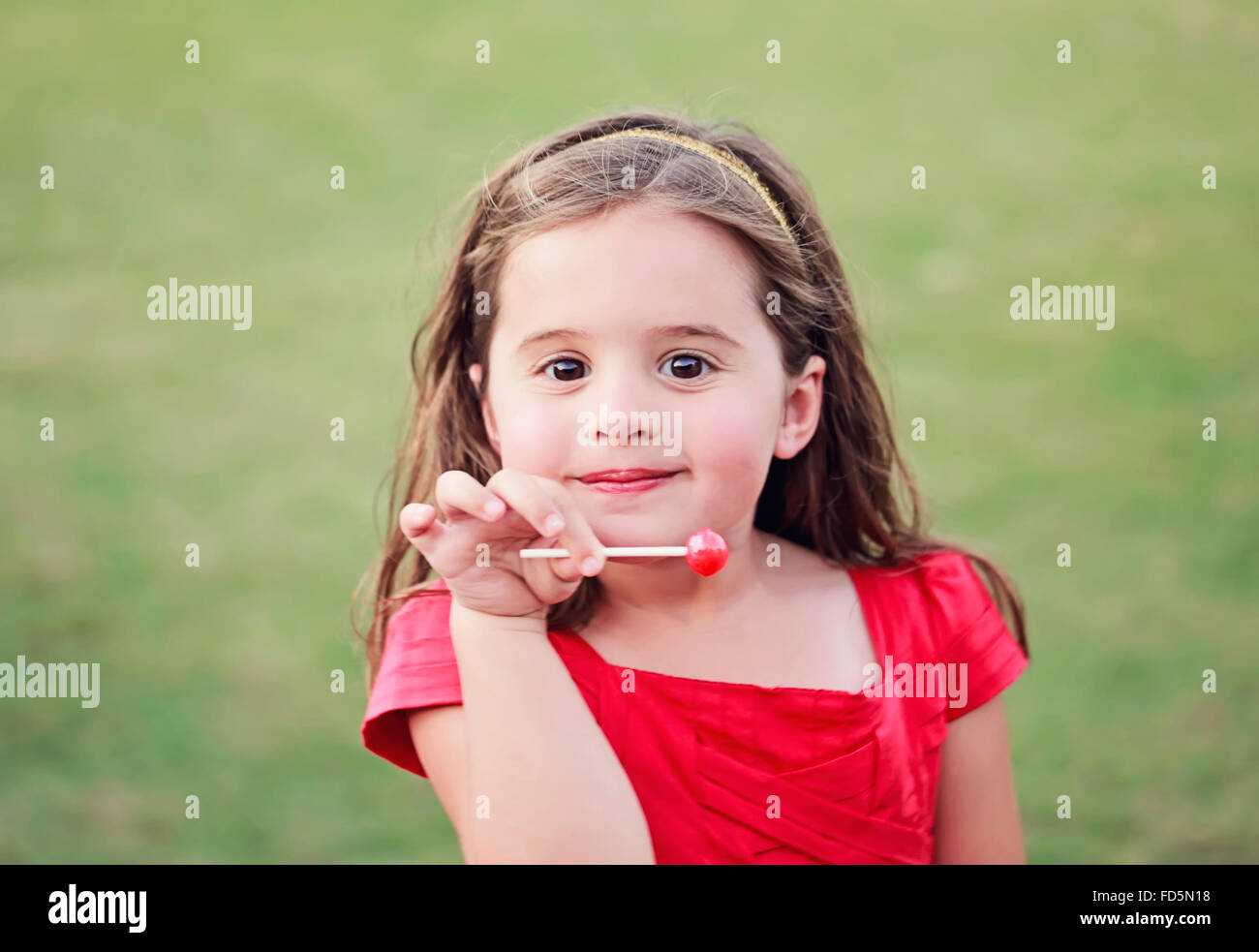 Little girl in a bright red dress holding a bright red lollipop while smiling at the camera. Stock Photo