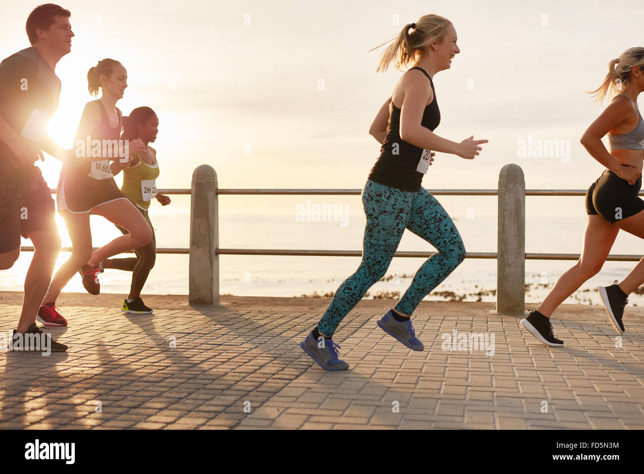 Portrait of young people running on seaside promenade. Men and women running marathon on road by the sea at sunset. Stock Photo