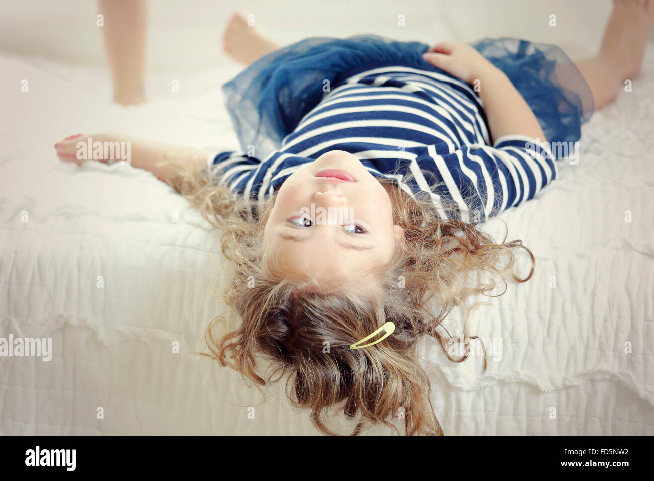 Image of a little girl laying on her bed with her head hanging off the edge so that she's upside down. Stock Photo