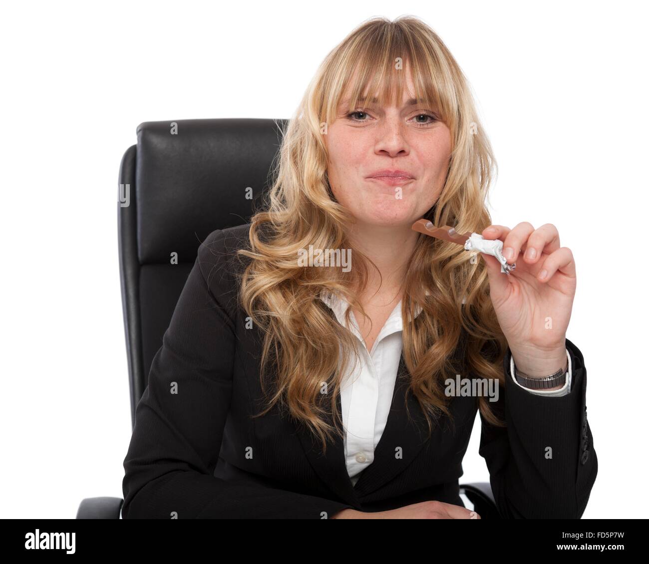 Beautiful young blond businesswoman eating a candy at her desk looking up at the camera with a mouthful and a guilty smile Stock Photo
