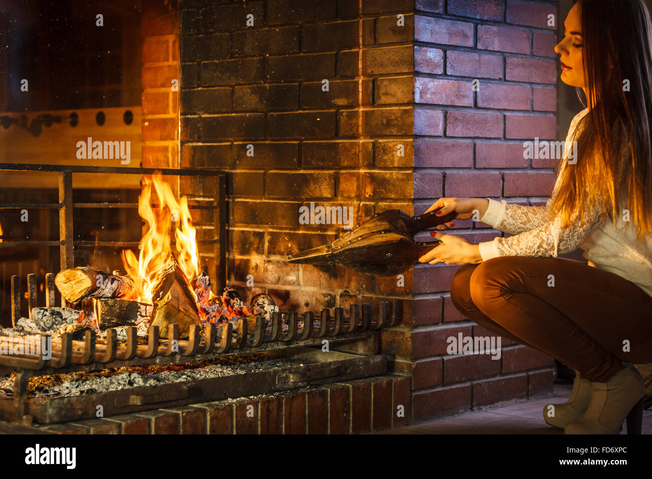 Woman at fireplace making fire with bellows. Young girl heating warming up and relaxing. Winter at home. Stock Photo