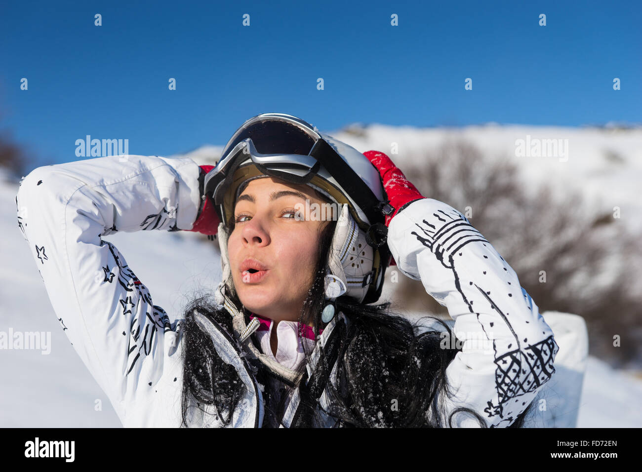 Head and Shoulders Close Up Playful Young Woman Wearing Helmet and Ski Goggles Standing on Snowy Mountainside with Hands Behind Head Enjoying Warm Sunshine on Beautiful Day. Stock Photo