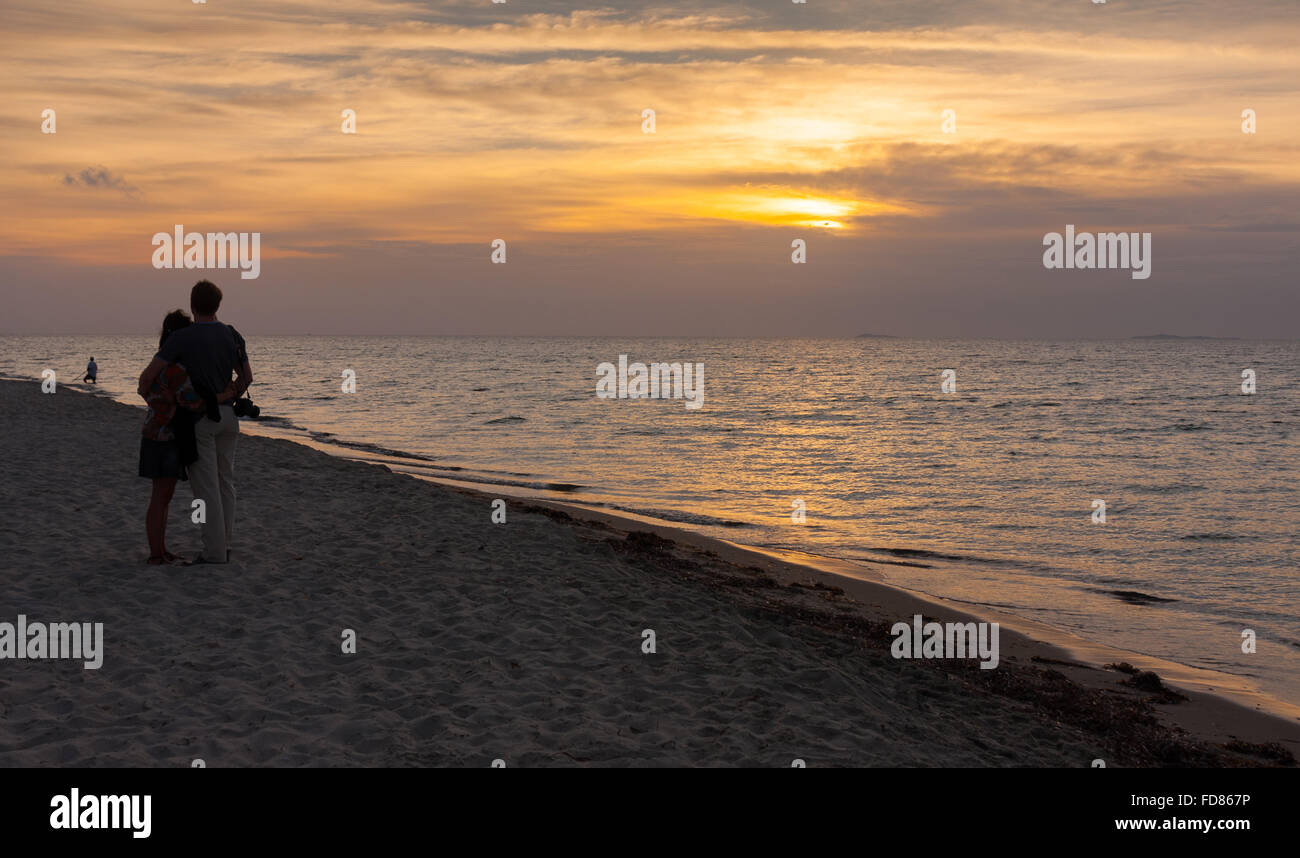 couple in silhouette standing close together on beach watching cloudy sunset Stock Photo