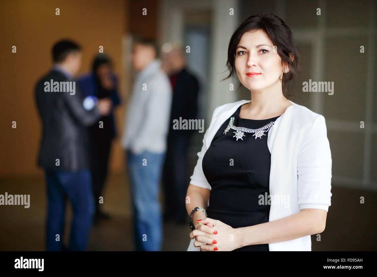 Successful attractive business woman boss brunette with kind eyes stands inside office building and welcoming smile. Strict black dress jewelry. Cute young chief female posing. In background man discussing. Stock Photo