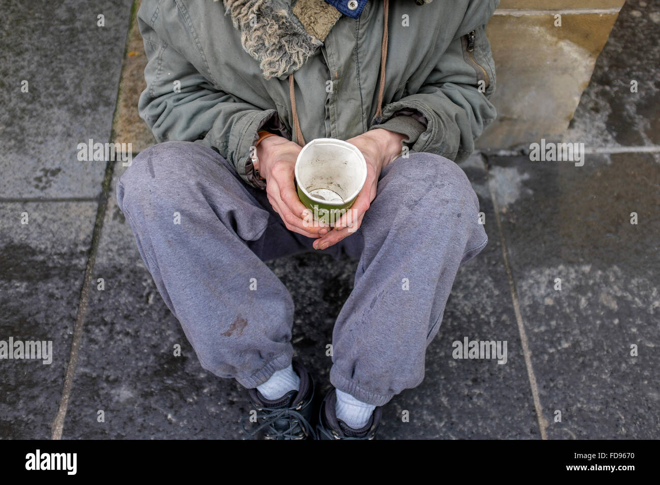 Street beggar, sitting on a pavement with a cardboard cup for collecting money and charity. Stock Photo