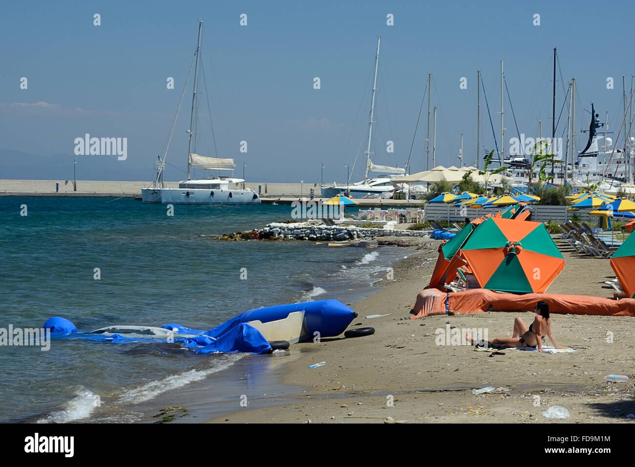 Collapsed inflatable dinghy used by migrants to reach Greece from Turkey on the beach at Kos Town with a woman tourist sunbathin Stock Photo