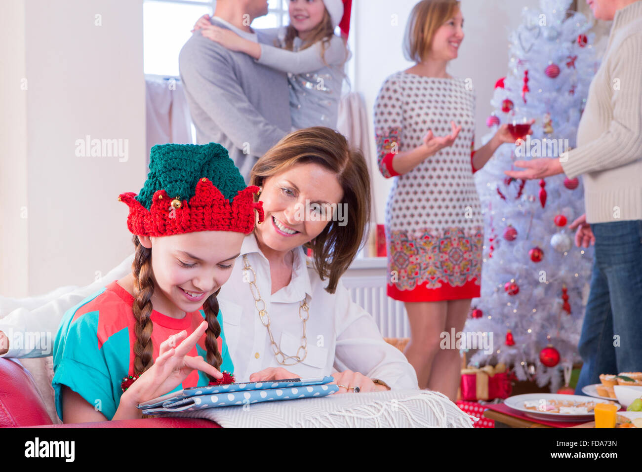 Three Generation Family at Christmas Time. A young girl and her grandma focus on the tablet while the rest of the family sociali Stock Photo