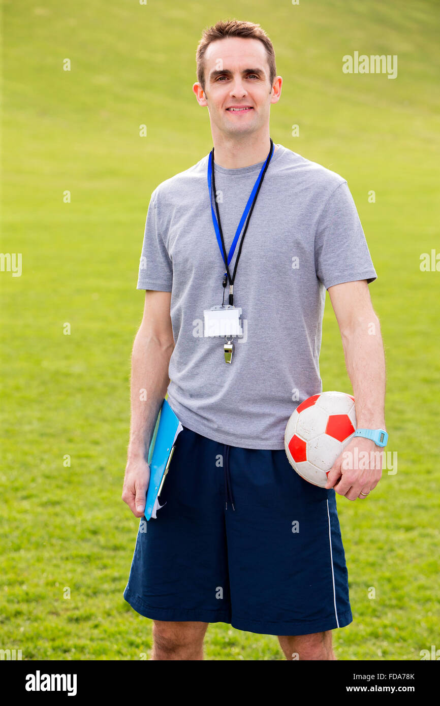 A male sports teacher stands outdoors in a field, he is holding a clipboard and football Stock Photo