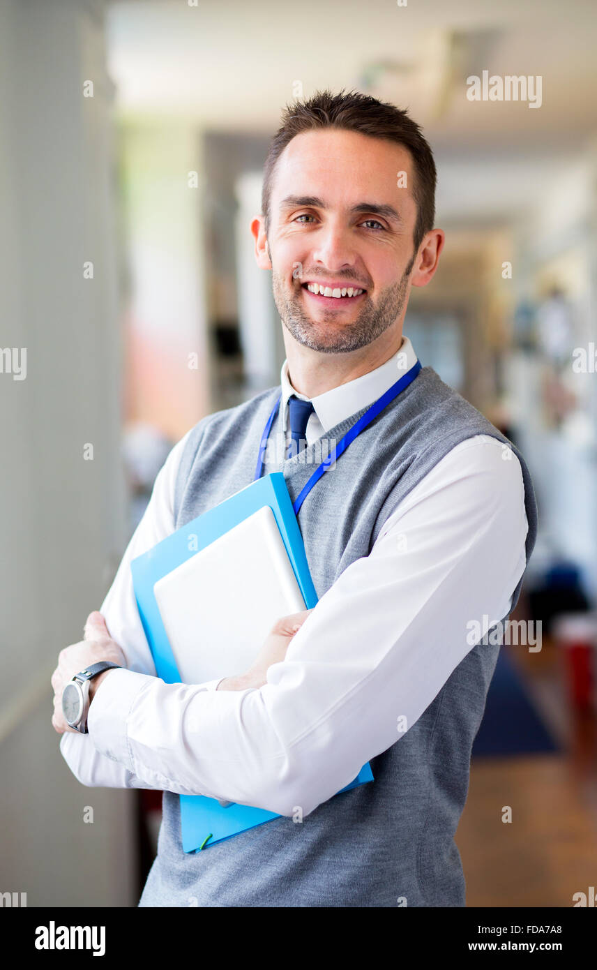 A happy male teacher dressed smartly and smiling in a school corridor. He is holding folders and a digital tablet. Stock Photo