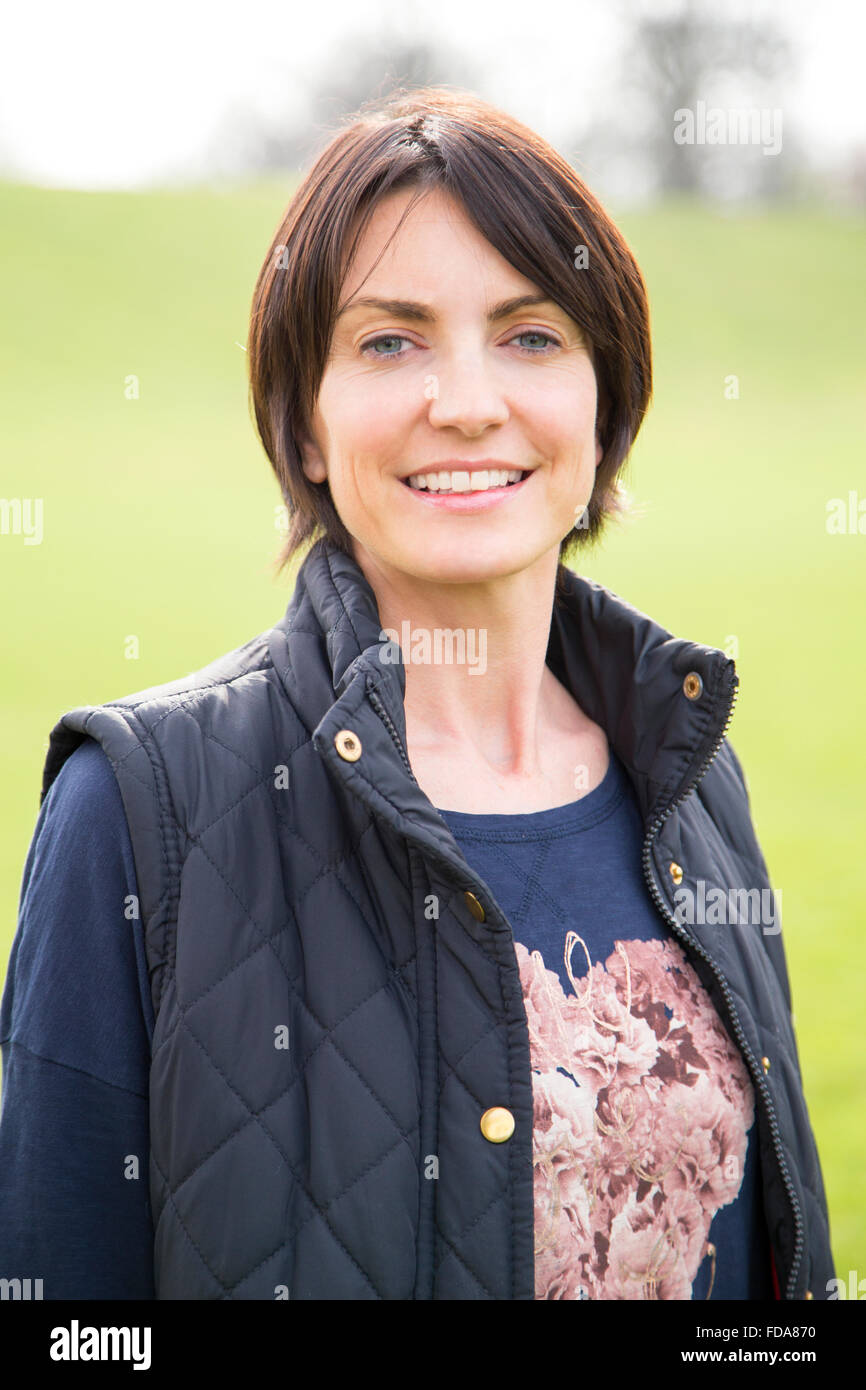 Head and shoulder portrait of a woman looking at the camera, she is outdoors. A grassy area can be seen behind her, She smiles. Stock Photo