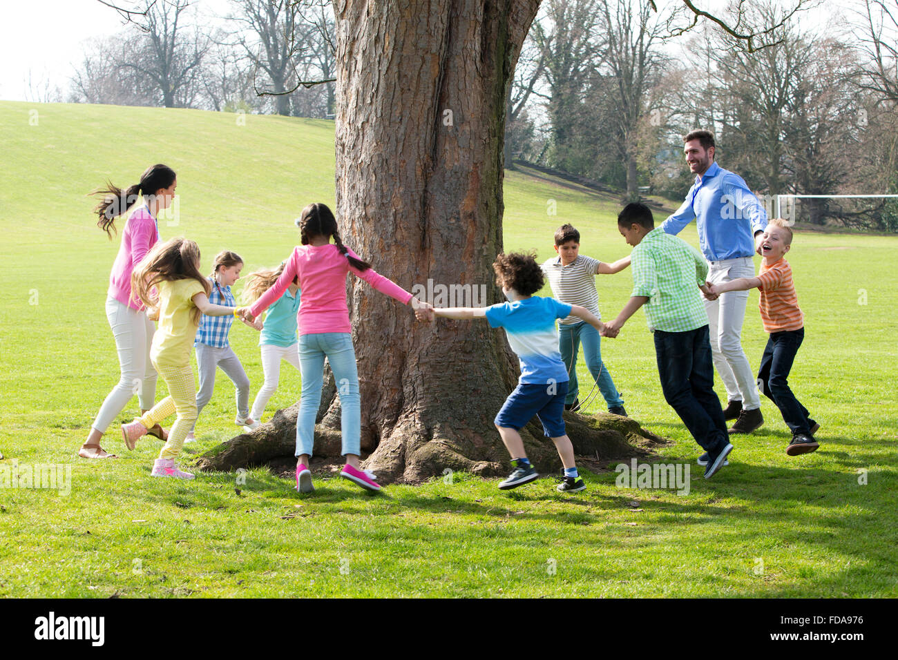 A group of children with two adults holding hands and dance around the trunk of a tree. Stock Photo