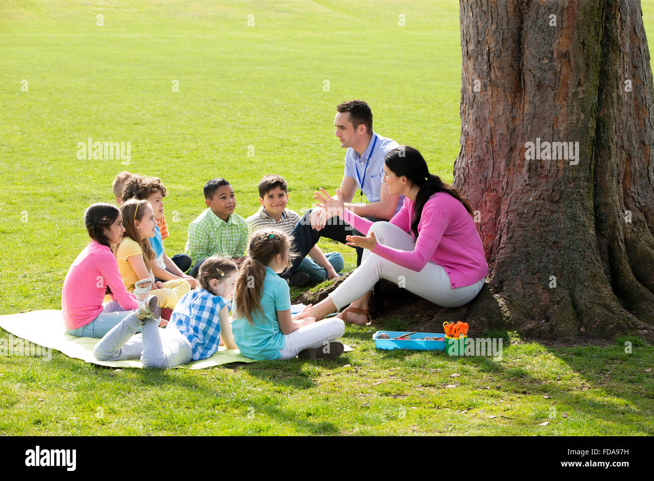 Small group of children sitting on the grass having a lesson outdoors. Male and female teacher can be seen. The children look to Stock Photo