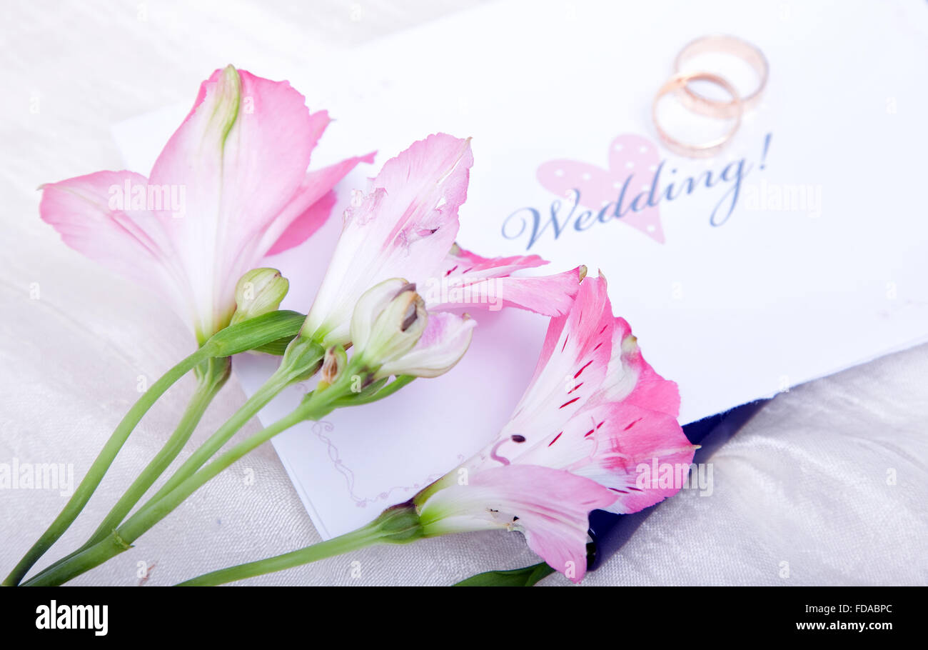 Beautiful pink lilies rest on top of a wedding invitation with the wedding rings out of focus in the background. Stock Photo