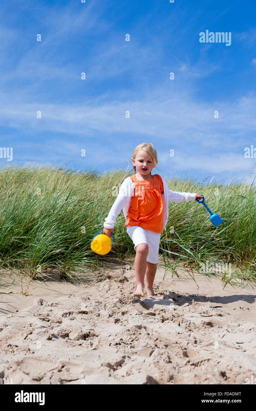 Little Girl running onto the beach with her arms outstretched and holding a bucket and spade. Stock Photo