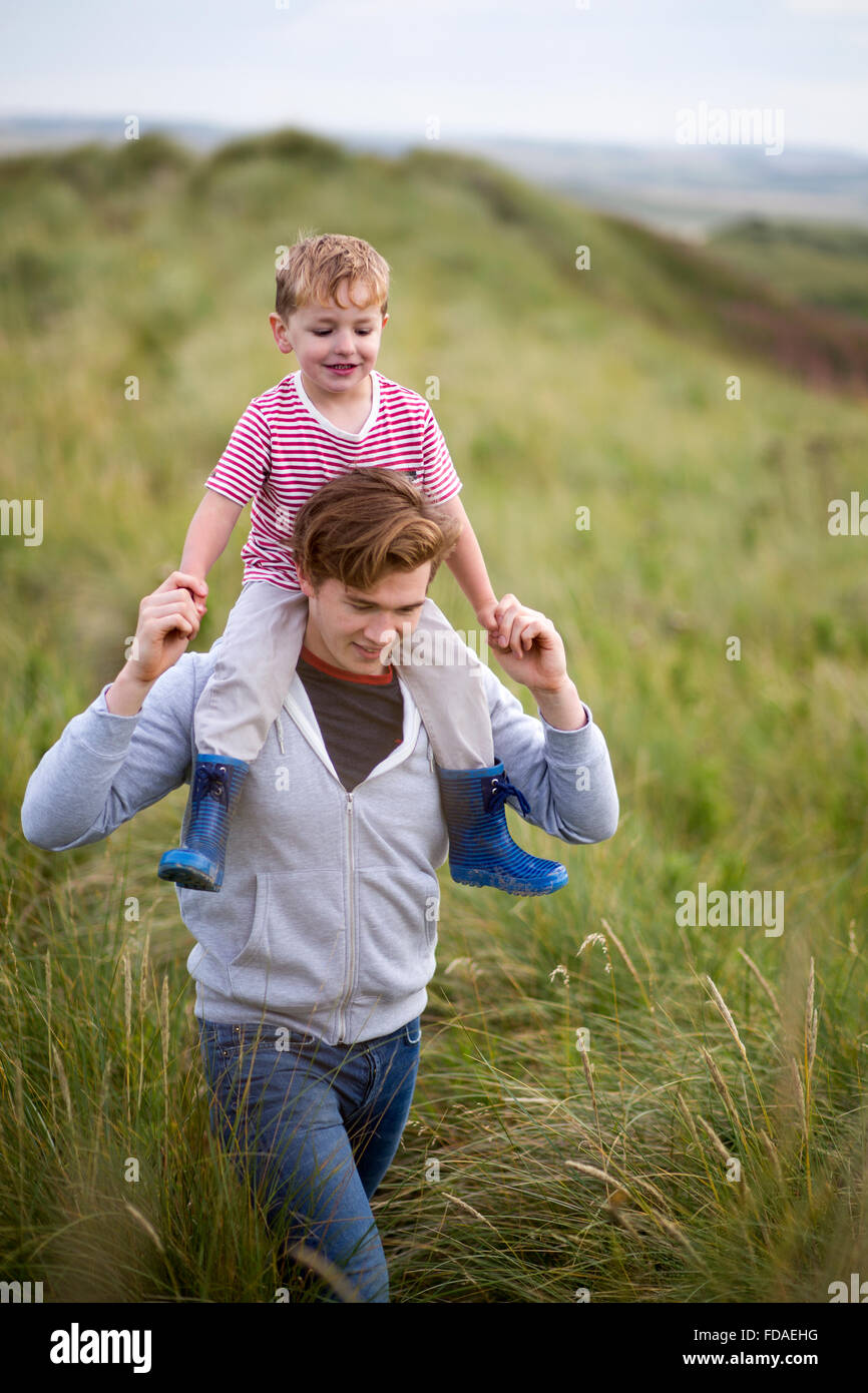 Portrait of a father and son walking through long grass next to a beach. The little boy is on his fathers shoulders and they are Stock Photo