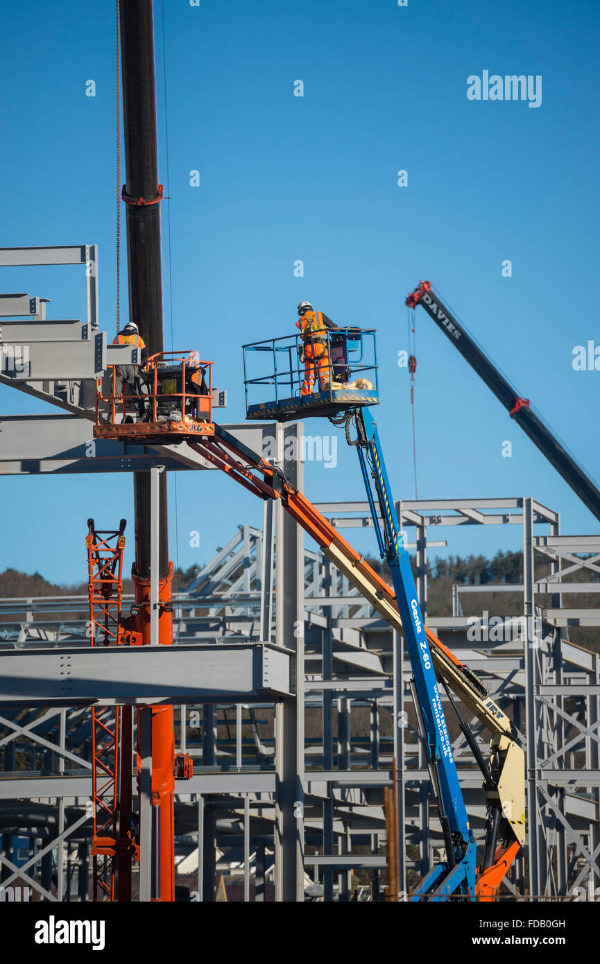 Men working at height from 'cherry picker' elevated platforms - erecting bolting together the steel frame of a new building that will house a branch of Tesco supermarket and Marks & Spencer store,  on a clear blue sky day, Aberystwyth Wales UK Stock Photo