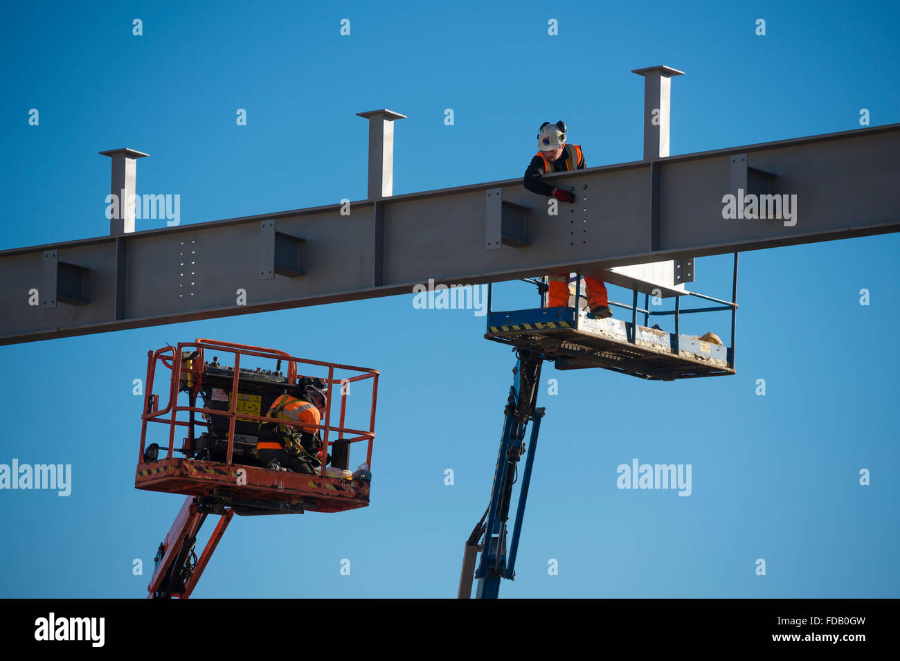 Men working at height from 'cherry picker' elevated platforms - erecting bolting together the steel frame of a new building that will house a branch of Tesco supermarket and Marks & Spencer store,  on a clear blue sky day, Aberystwyth Wales UK Stock Photo