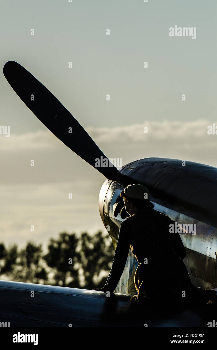 A girl in period costume sits on the wing of a wartime P-40 Kittyhawk at the Goodwood Revival 2015, in silhouette Stock Photo