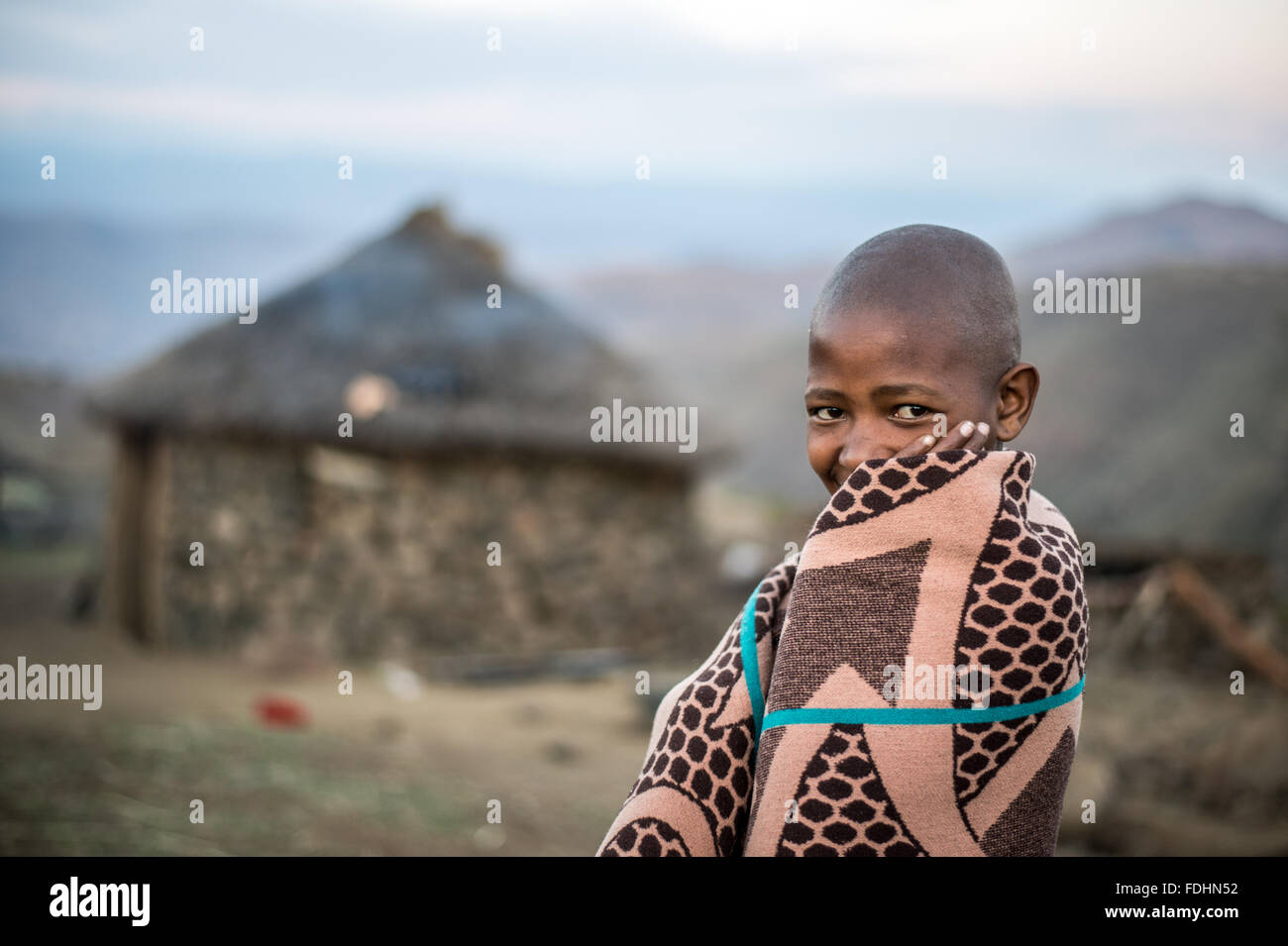 Young boy wrapped in a blanket standing in front of a stone hut in Lesotho, Africa Stock Photo