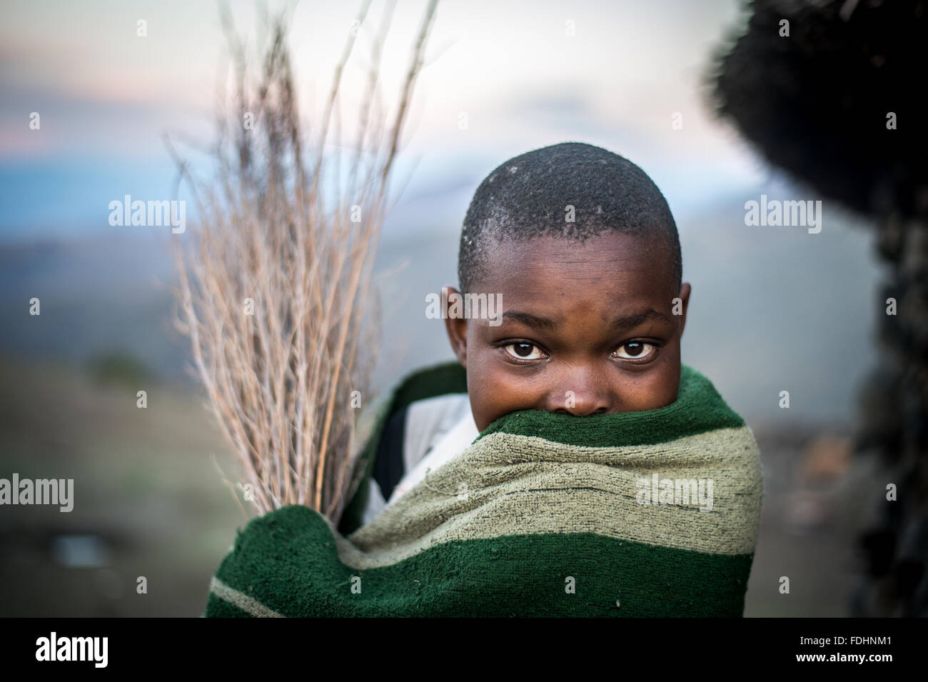 Young boy wrapped in a blanket standing in front of a stone hut in Lesotho, Africa Stock Photo