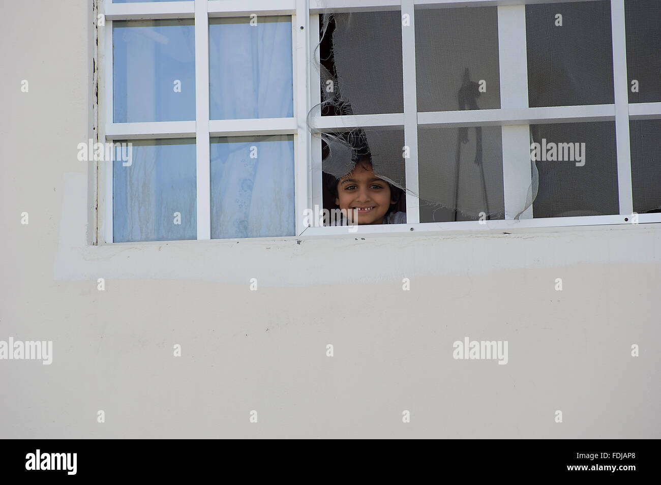 A child in Muscat, Oman smiling with a gap in his teeth and looking out of a broken window through a torn screen Stock Photo