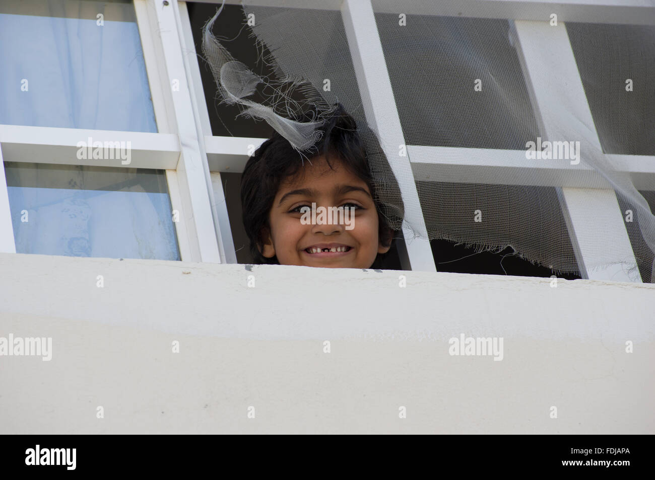 A child in Muscat, Oman smiling with a gap in his teeth and looking out of a broken window through a torn screen Stock Photo
