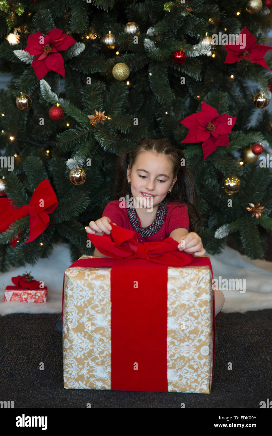 Smiling Girl sitting by Christmas tree opening gift Stock Photo