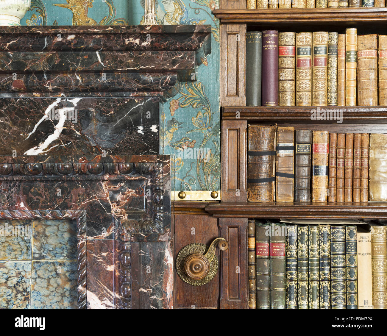 Detail of a corner of the fireplace, a part of the bookshelves and the Victorian Cordelova embossed wallpaper in the Library at Dunster Castle, Somerset. Stock Photo