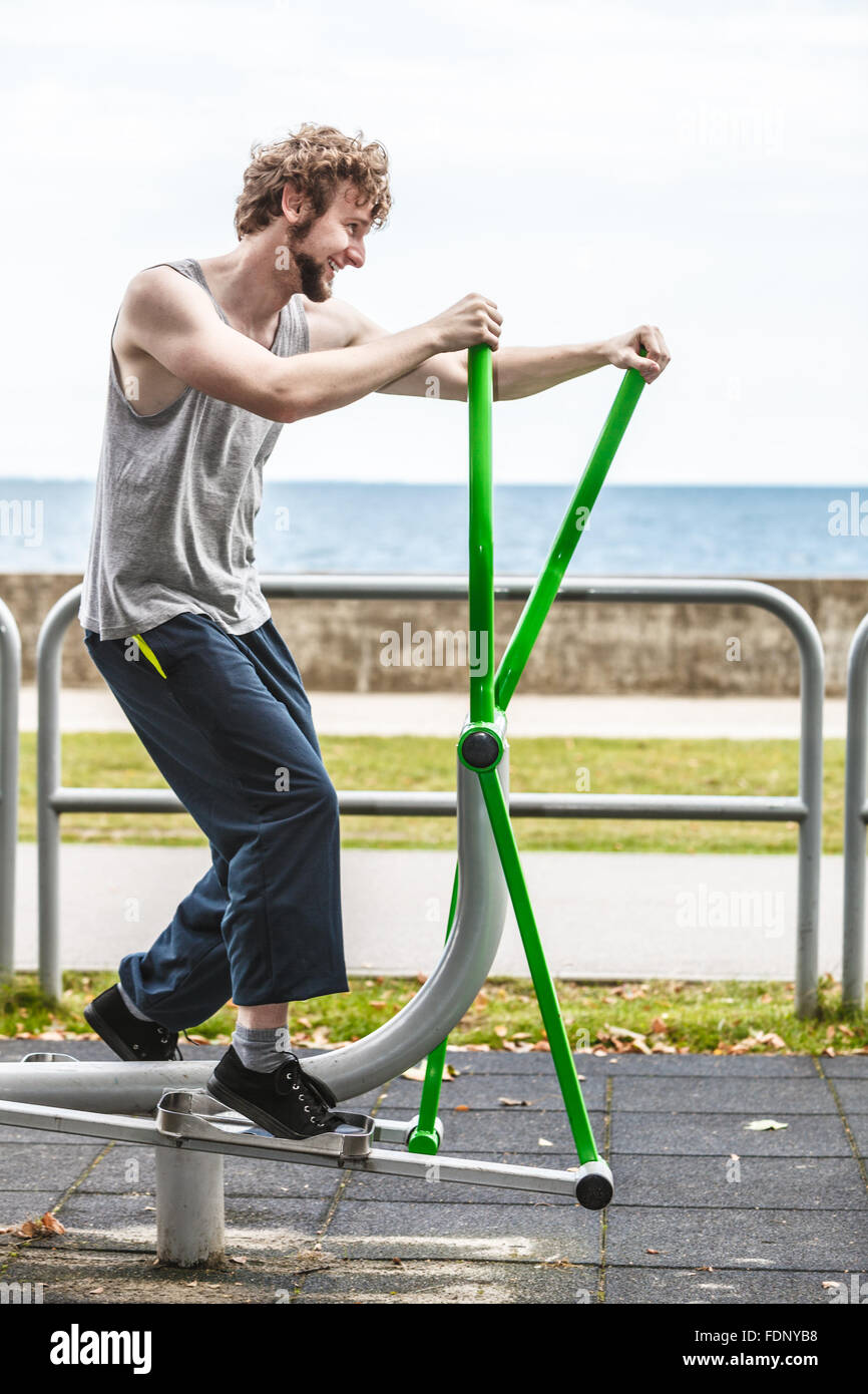 Active young man exercising on elliptical trainer machine. Muscular sporty guy in training suit working out at outdoor gym. Sport fitness and healthy lifestyle concept. Stock Photo