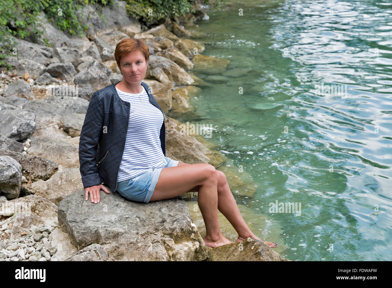 white middle aged red haired tanned woman in shorts and leather jacket sits barefoot on stones by the lake shore with clear tran Stock Photo