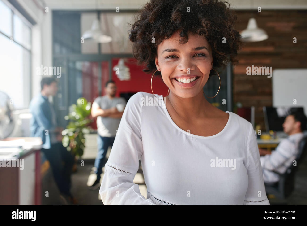 Portrait of smiling young african businesswoman with people in background. Cheerful young woman standing relaxed in her office, Stock Photo