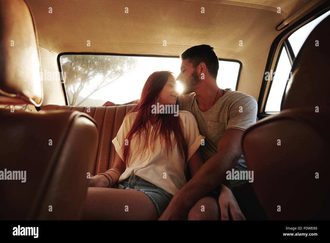 Affectionate young couple sitting in back seat of a car. Young man and woman in rear seat of a vehicle with sun flare. Stock Photo