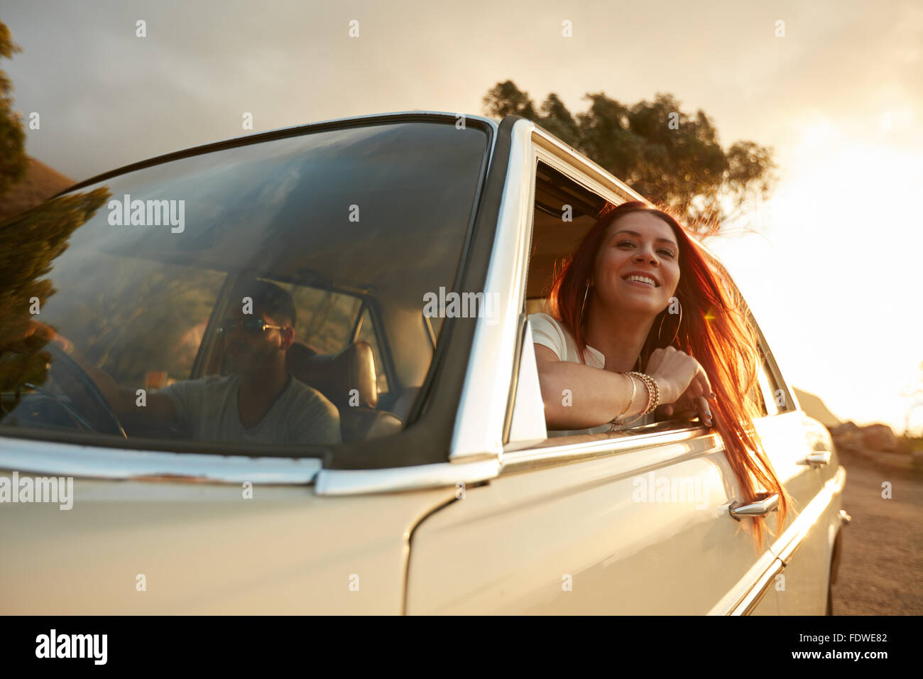 Portrait of happy young woman going on a road trip leaning out of window. Female enjoying travelling in a car with her boyfriend Stock Photo