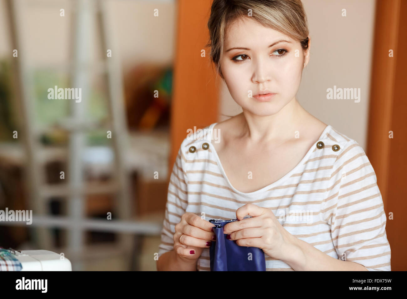 Young woman sewing at home, hemming blue fabric, stands and looks pensively to the side. Copy space. Fashion designer creating new fashionable styles. Stock Photo