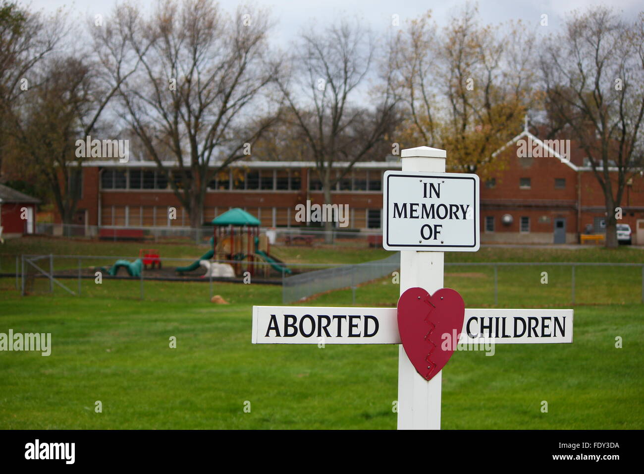 White wooden cross with heart in memory of aborted children in front of empty playground Stock Photo