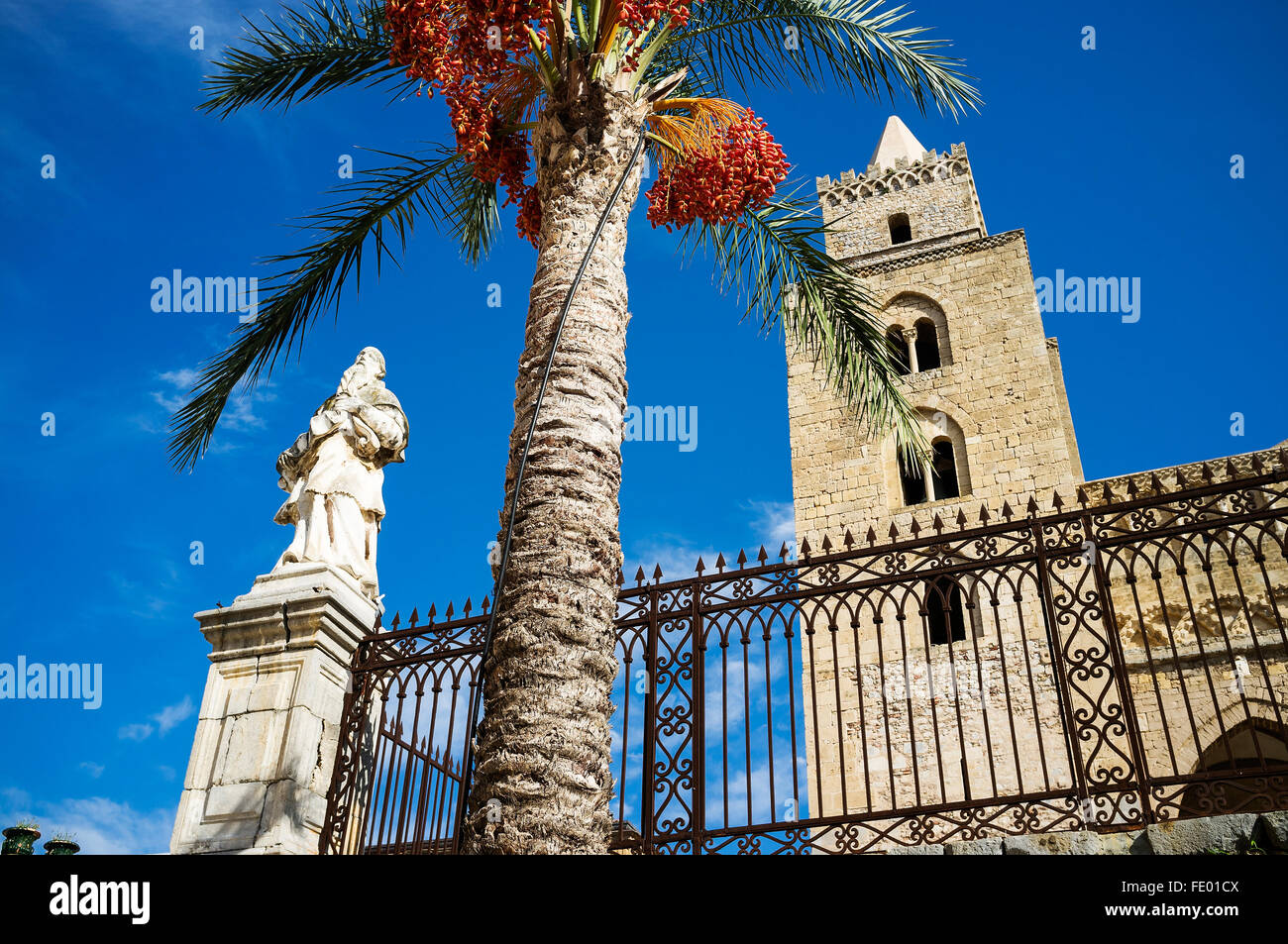 Low angle view of church tower and palm tree with religious statue in Cefalu city and comune in the Province of Palermo, Sicily, Stock Photo