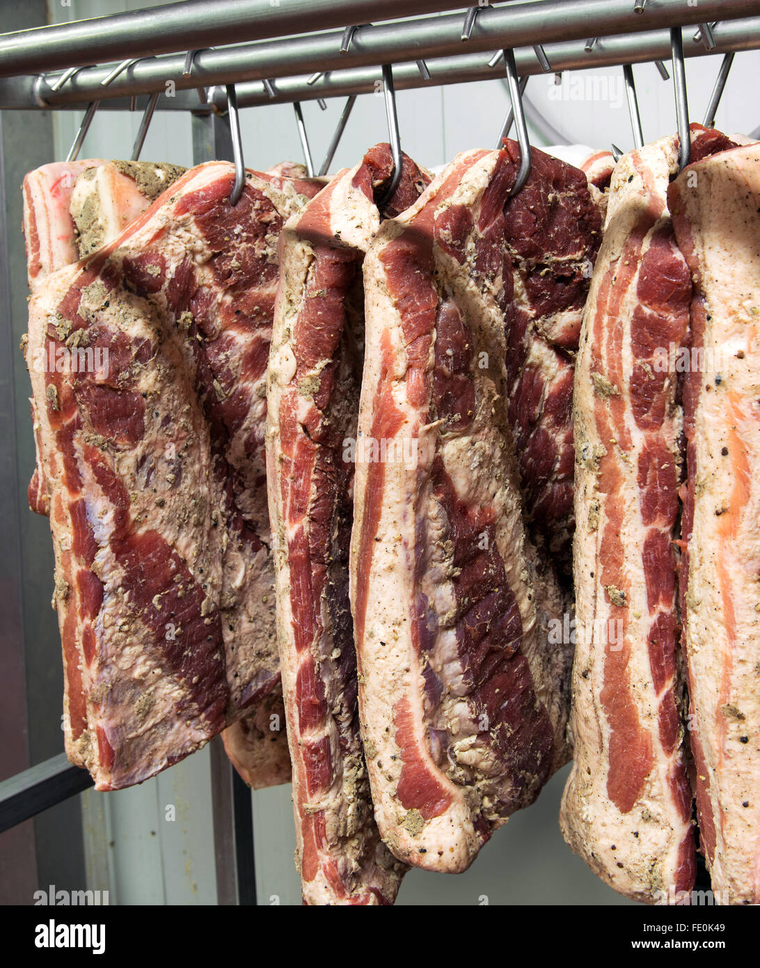 Multiple slabs of freshly processed meat covered with seasoning hanging on hooks in food processing factory Stock Photo