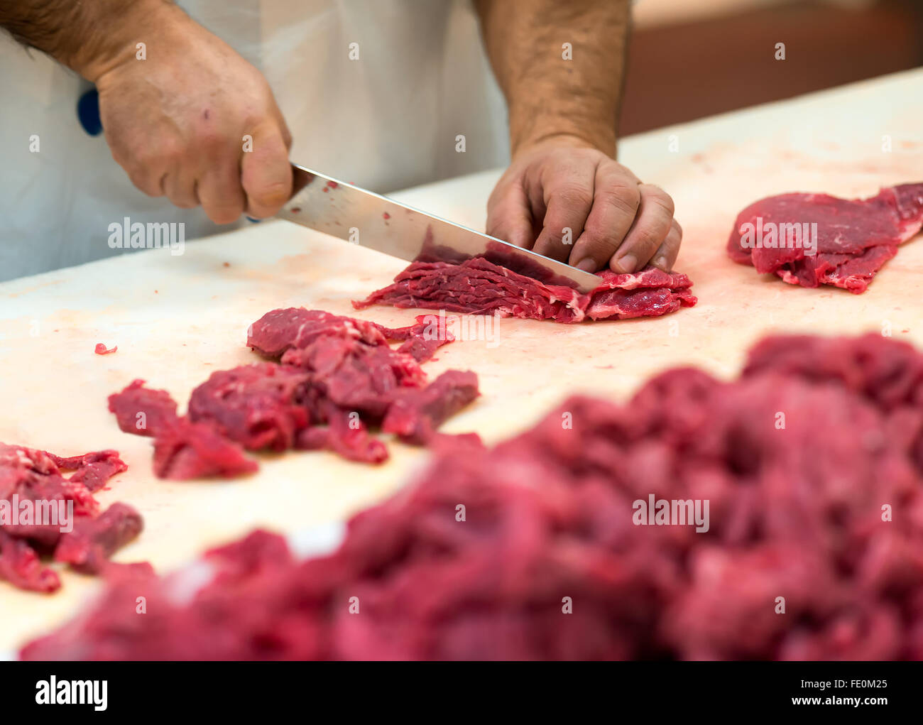 Close up of hands holding butcher knife and cuts of raw red meat on long cutting board in food processing facility Stock Photo