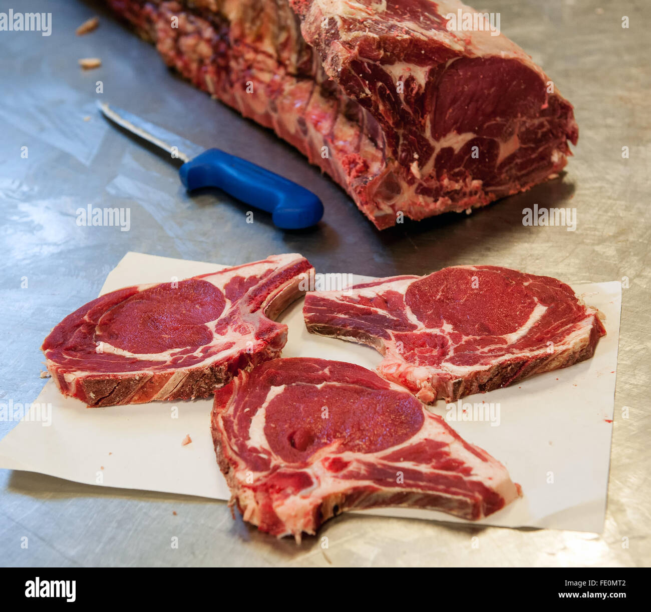 Three slices of raw rib eye steak over butcher paper sheet next to knife and meat section on metal table Stock Photo